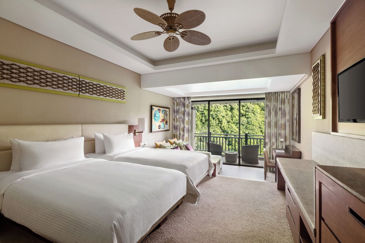 A hotel room with two beds, a ceiling fan, a balcony with a view of greenery, and a TV on the right side.