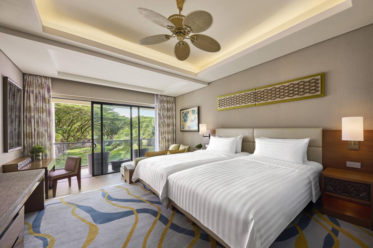 A hotel room with two beds, a desk, a chair, a balcony with a view of greenery, and a ceiling fan.