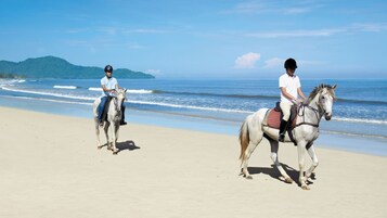Two individuals riding horses on a beach with ocean and hills in the background.
