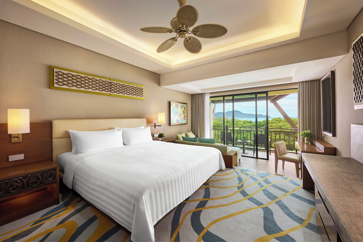 A hotel room with a large bed, a ceiling fan, a balcony with a view, and a desk with a chair.