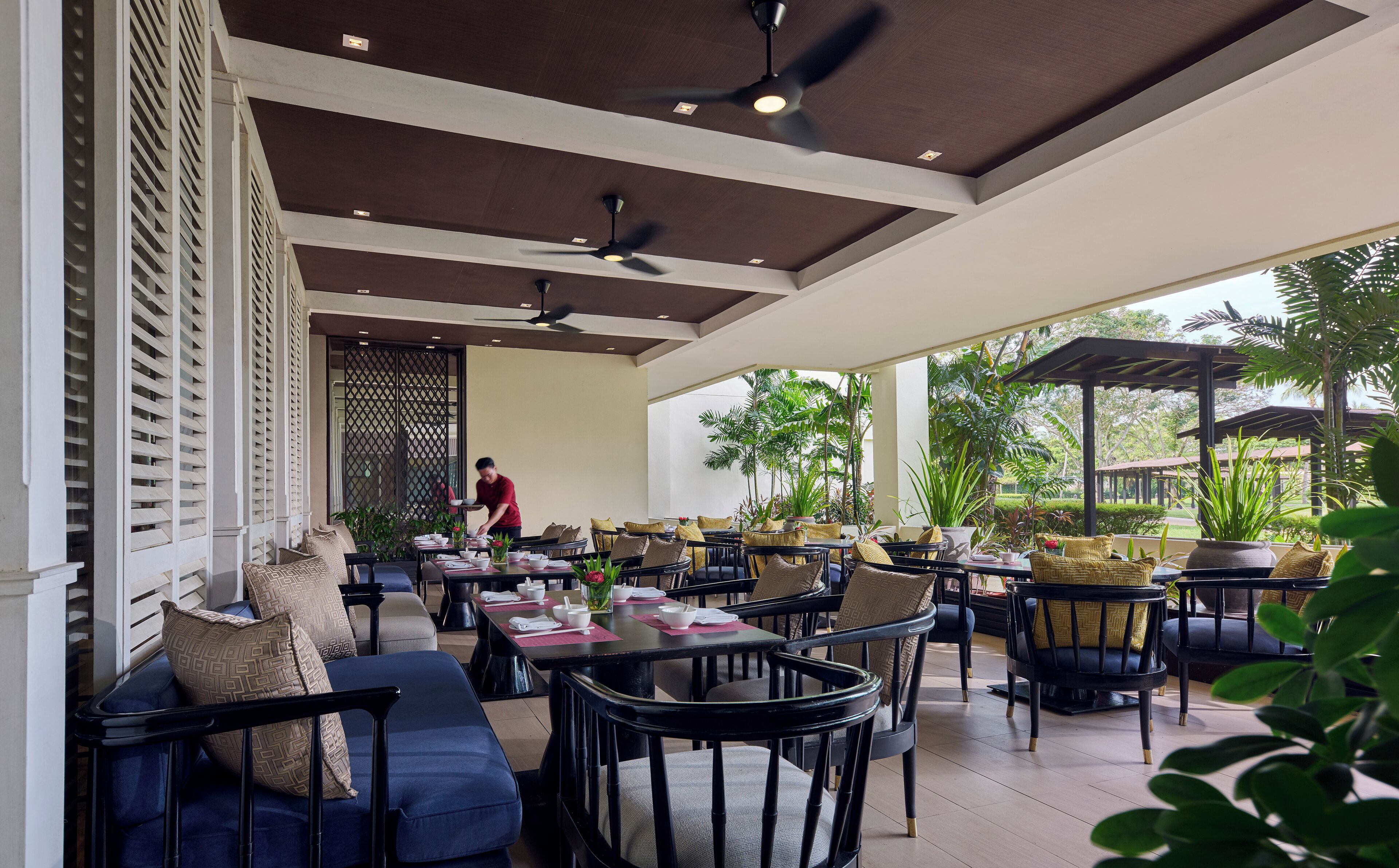 A covered outdoor dining area with tables and chairs, ceiling fans, and potted plants.