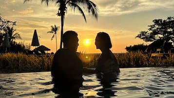 A couple sitting in a pool at sunset with palm trees and a house in the background.