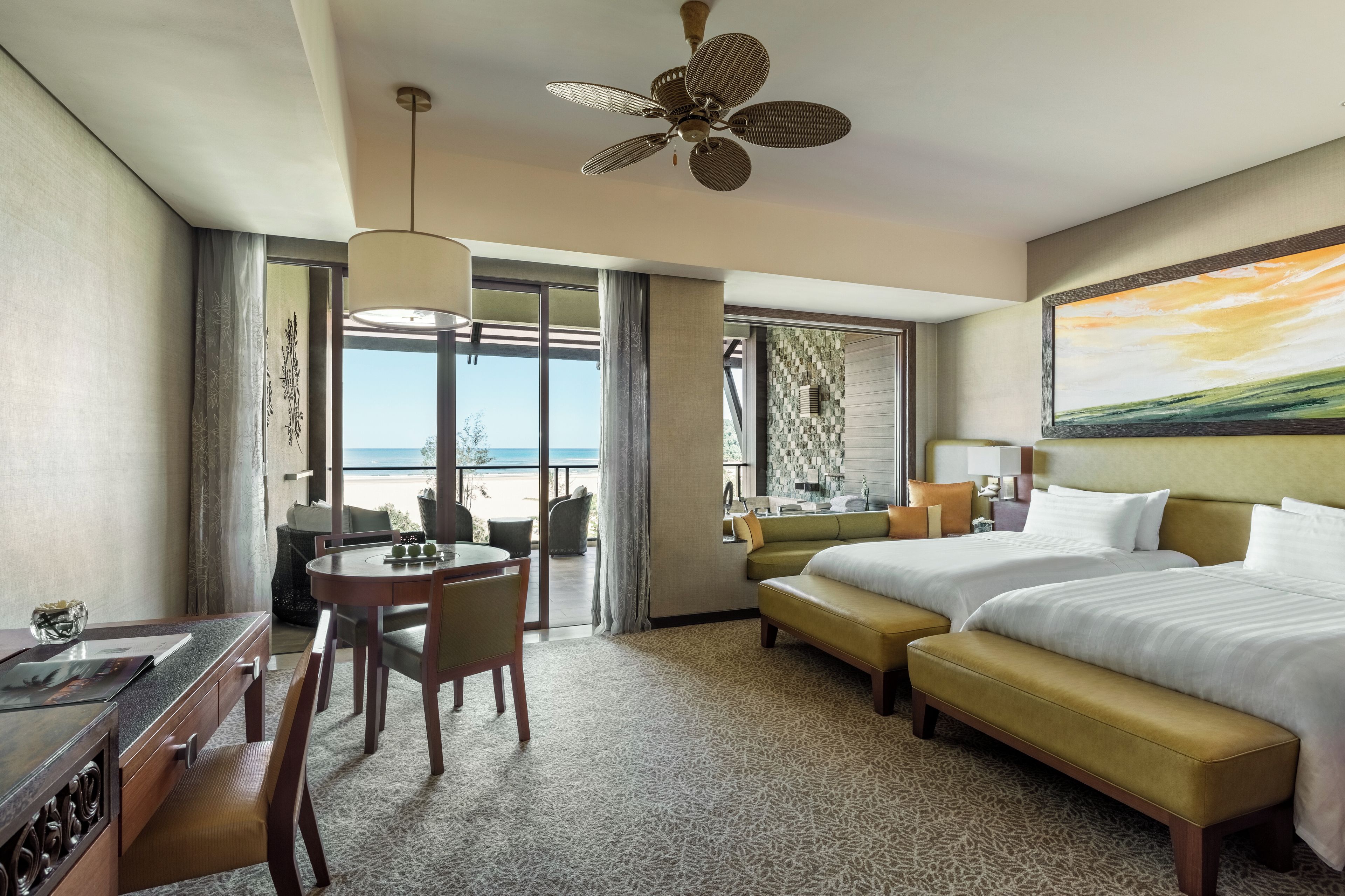 A hotel room with a bed, a desk with a chair, a dining table with chairs, a sofa, and a view of the beach.