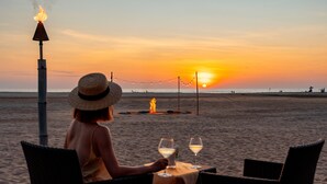 Escena de playa con una mesa, sillas, una fogata y un atardecer.