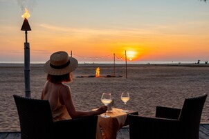 A beach scene with a table and chairs, a fire, and a sunset.
