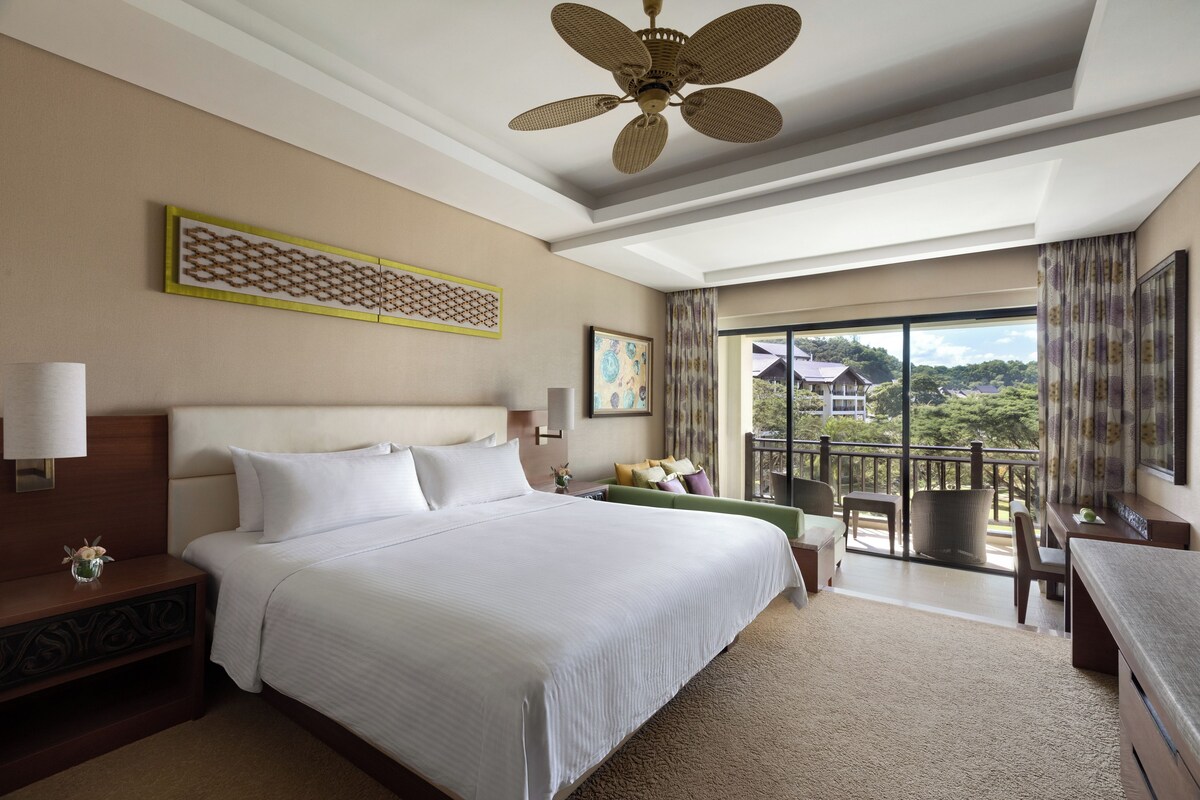 A hotel room with a large bed, a desk, a chair, a balcony with a view, and a ceiling fan.