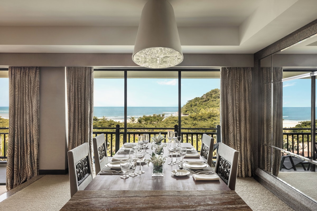 A dining area with a view of the ocean, large windows, and a chandelier.