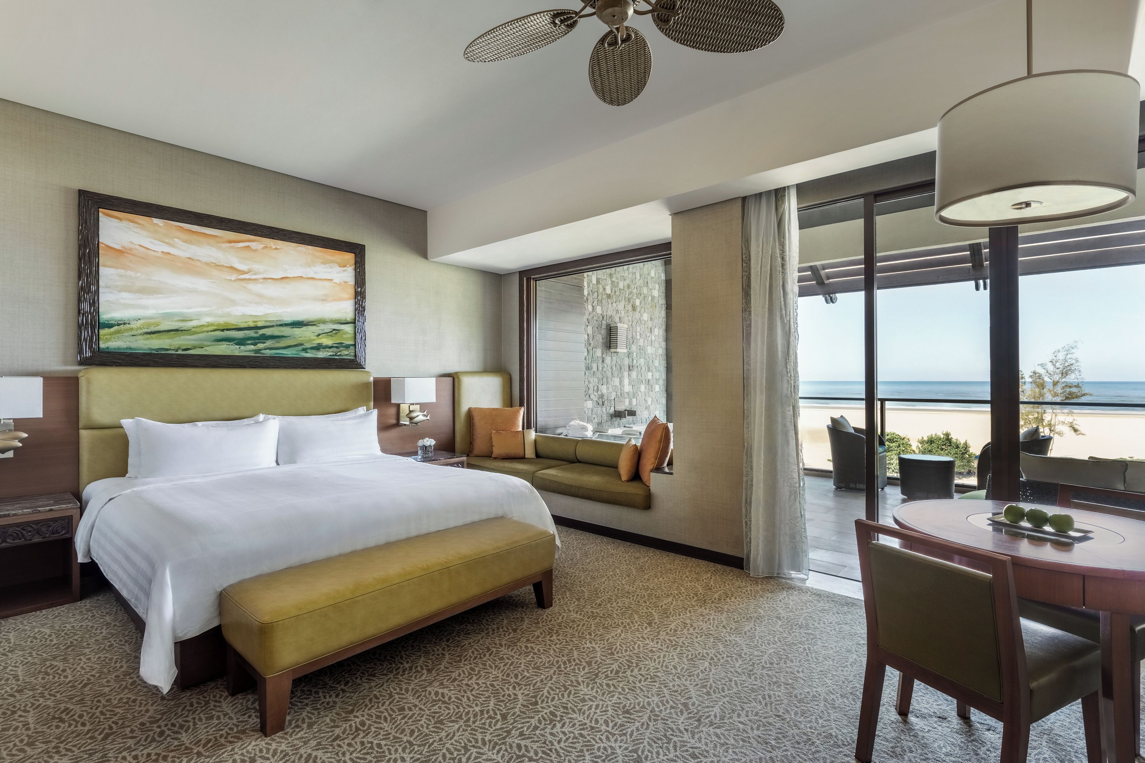 A hotel room with a bed, a sofa, a dining table with apples, and a view of the beach.