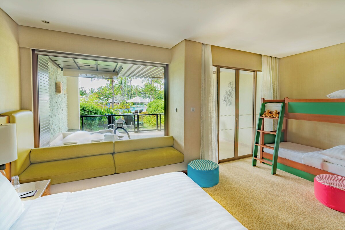 A hotel room with a bed, a bunk bed, a window with a view of a pool and greenery, and a small table with a lamp.