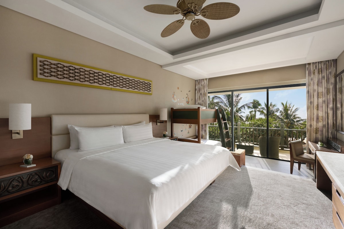 A hotel room with a large bed, a ceiling fan, a balcony with a view of palm trees, and a desk with a chair.