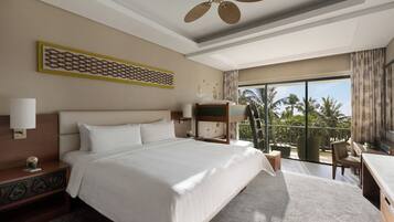 A hotel room with a large bed, a ceiling fan, a balcony with a view of palm trees, and a desk with a chair.