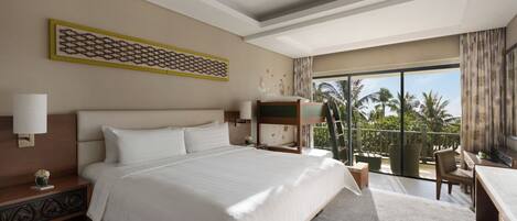 A hotel room with a large bed, a ceiling fan, a balcony with a view of palm trees, and a desk with a chair.