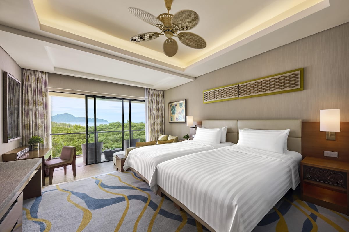 A hotel room with two beds, a desk, a chair, a large window with a view, and a ceiling fan.