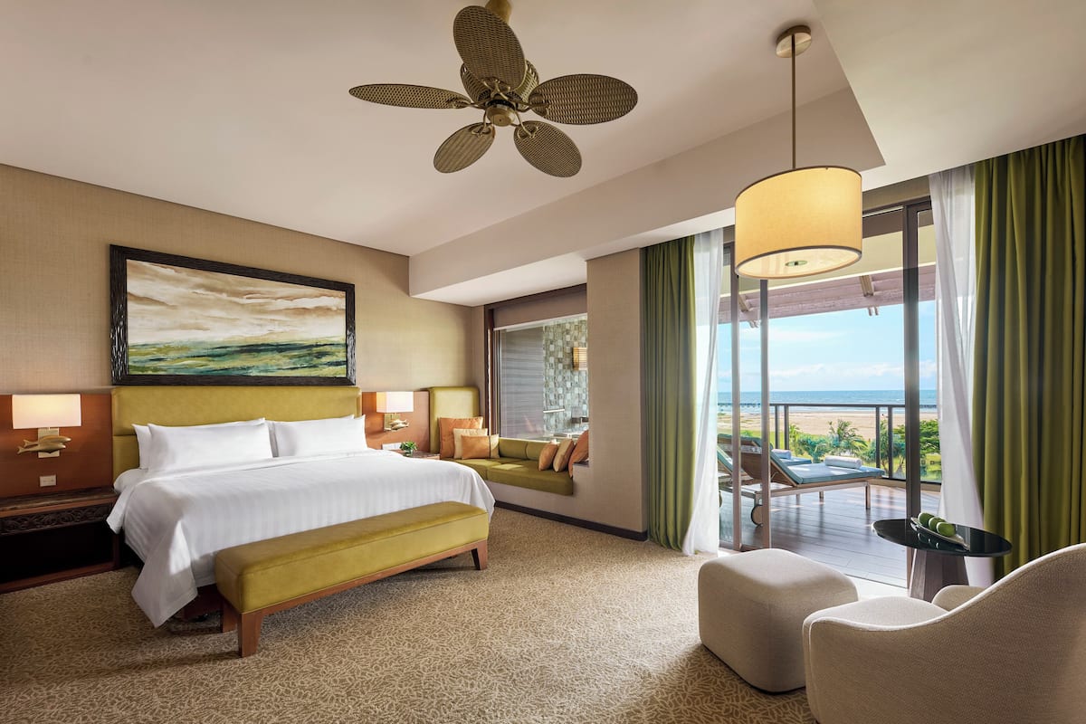 A hotel room with a large bed, a bench, a sofa, a balcony with a view of the beach, and a ceiling fan.