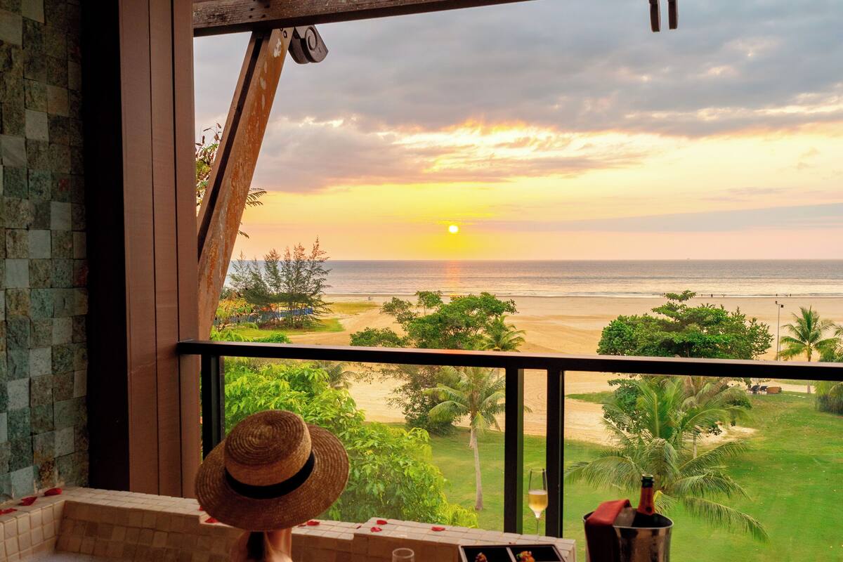 A person relaxing in a bathtub with a sunset view, a bottle of champagne, and rose petals on the floor.