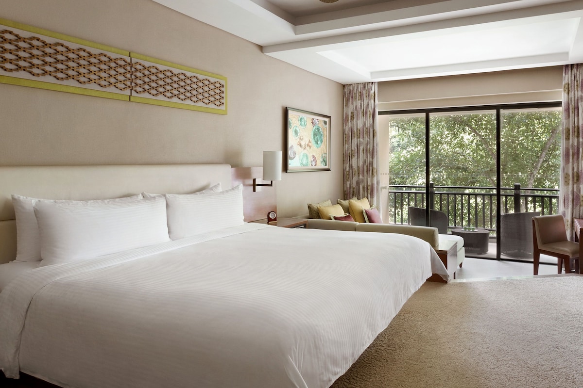 A large bed with white linens, a ceiling fan, a decorative wall piece, a painting, and a balcony with outdoor furniture.