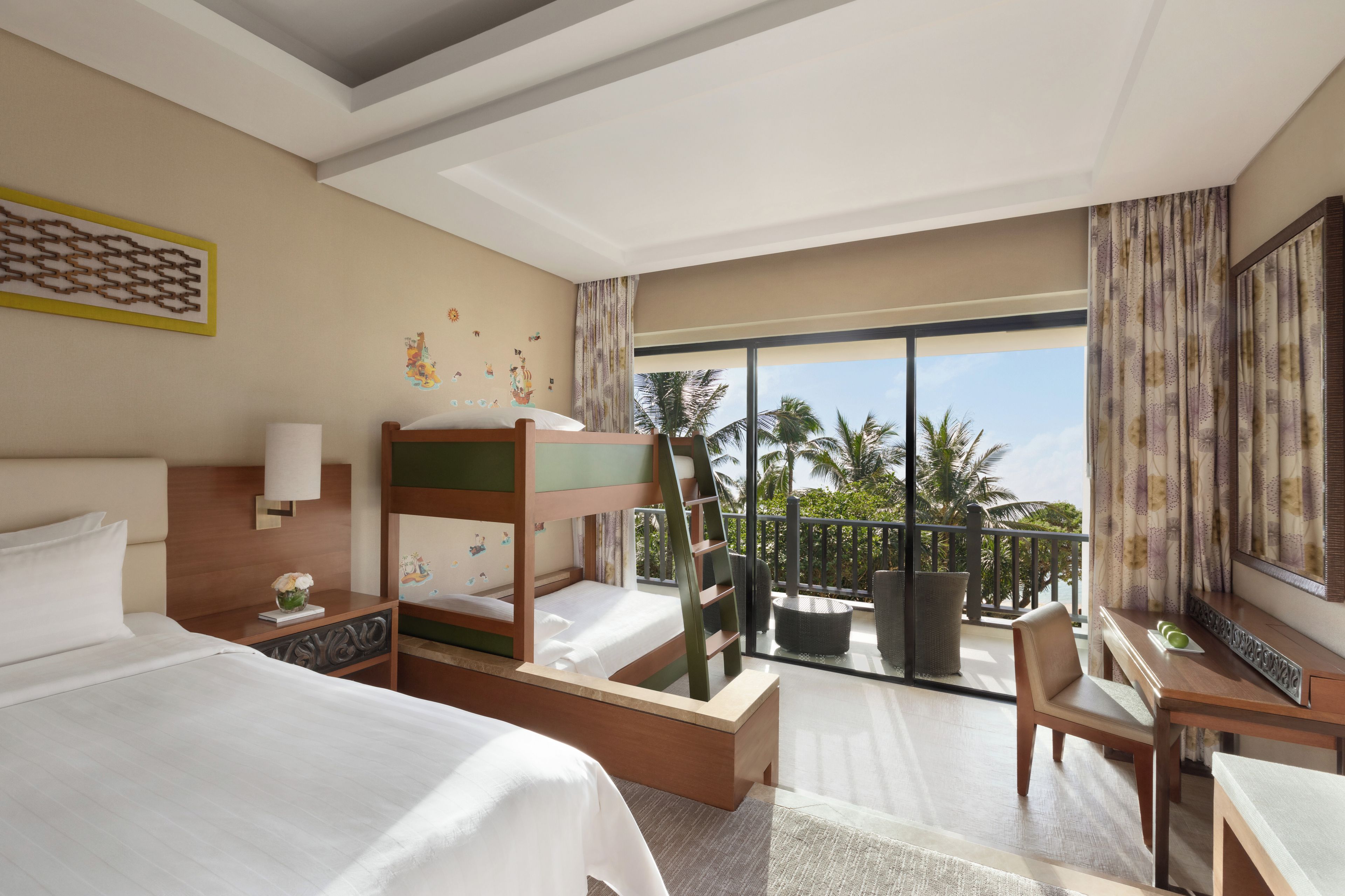 A hotel room with a bunk bed, a desk, a chair, a large window with a view of palm trees, and a balcony with a table and chairs.