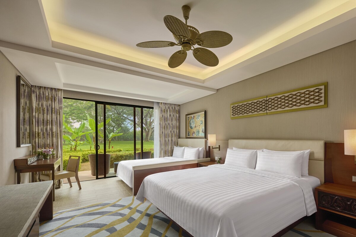 A hotel room with a large bed, two bedside tables, a desk, a chair, a ceiling fan, and a large window with a view of greenery.
