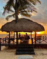 A thatched-roof outdoor dining area with a view of the sunset over the ocean.