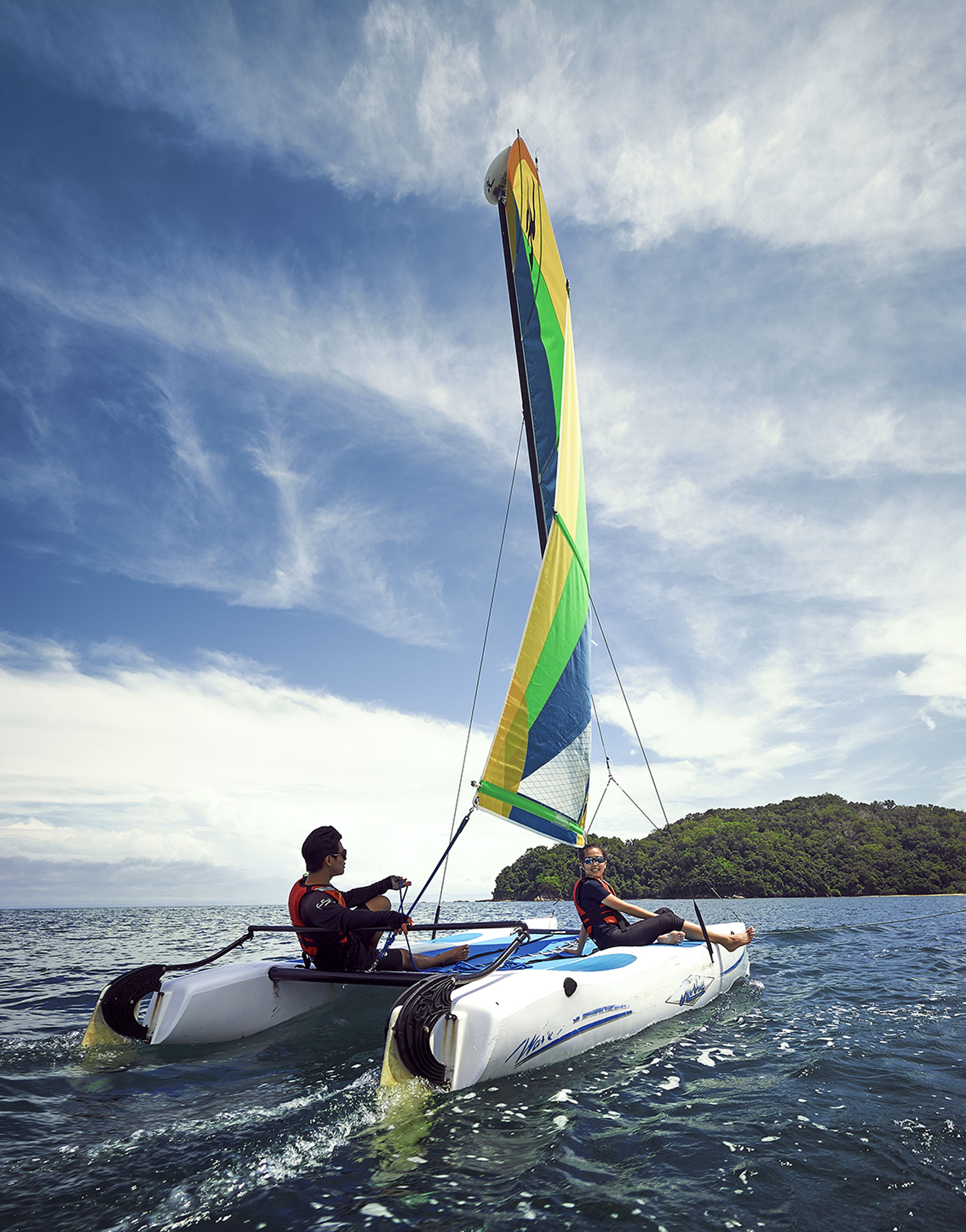 Un velero con dos personas a bordo, navegando sobre aguas tranquilas con una isla boscosa al fondo.