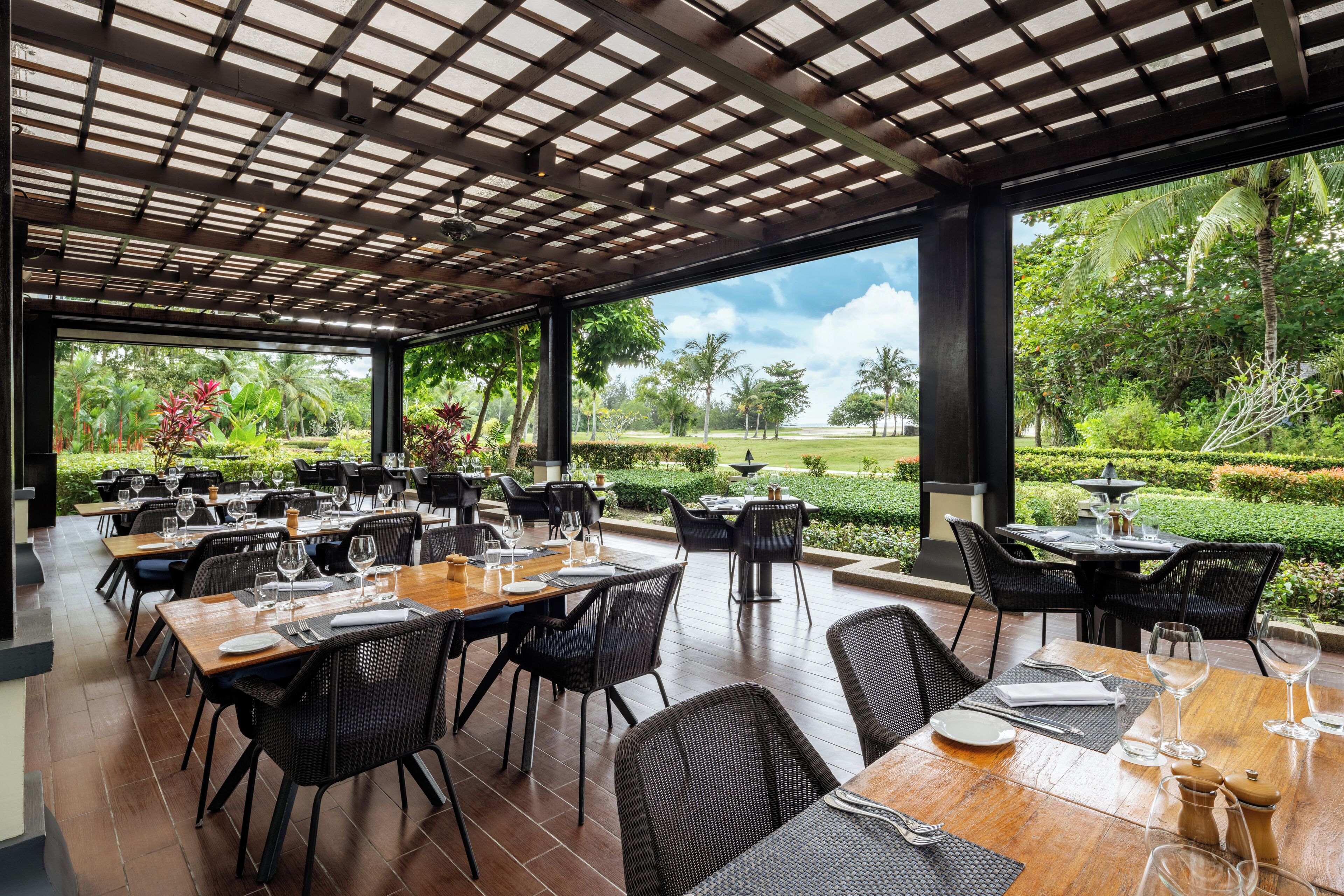 Un área de comedor al aire libre con mesas y sillas, una estructura de pérgola y vistas a un jardín paisajístico.
