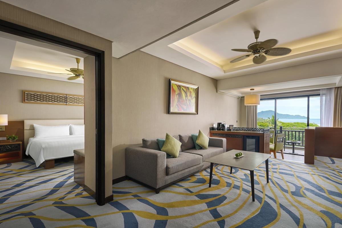 A hotel room with a bed, a sofa, a dining table, a balcony with a view, and a ceiling fan.