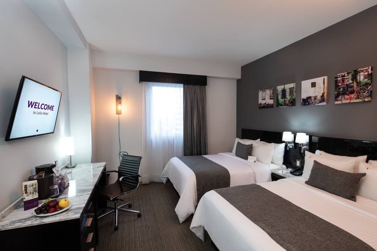 A hotel room with two beds, a desk with a chair, a TV displaying 'WELCOME to Laila Hotel', a window with curtains, and wall art.