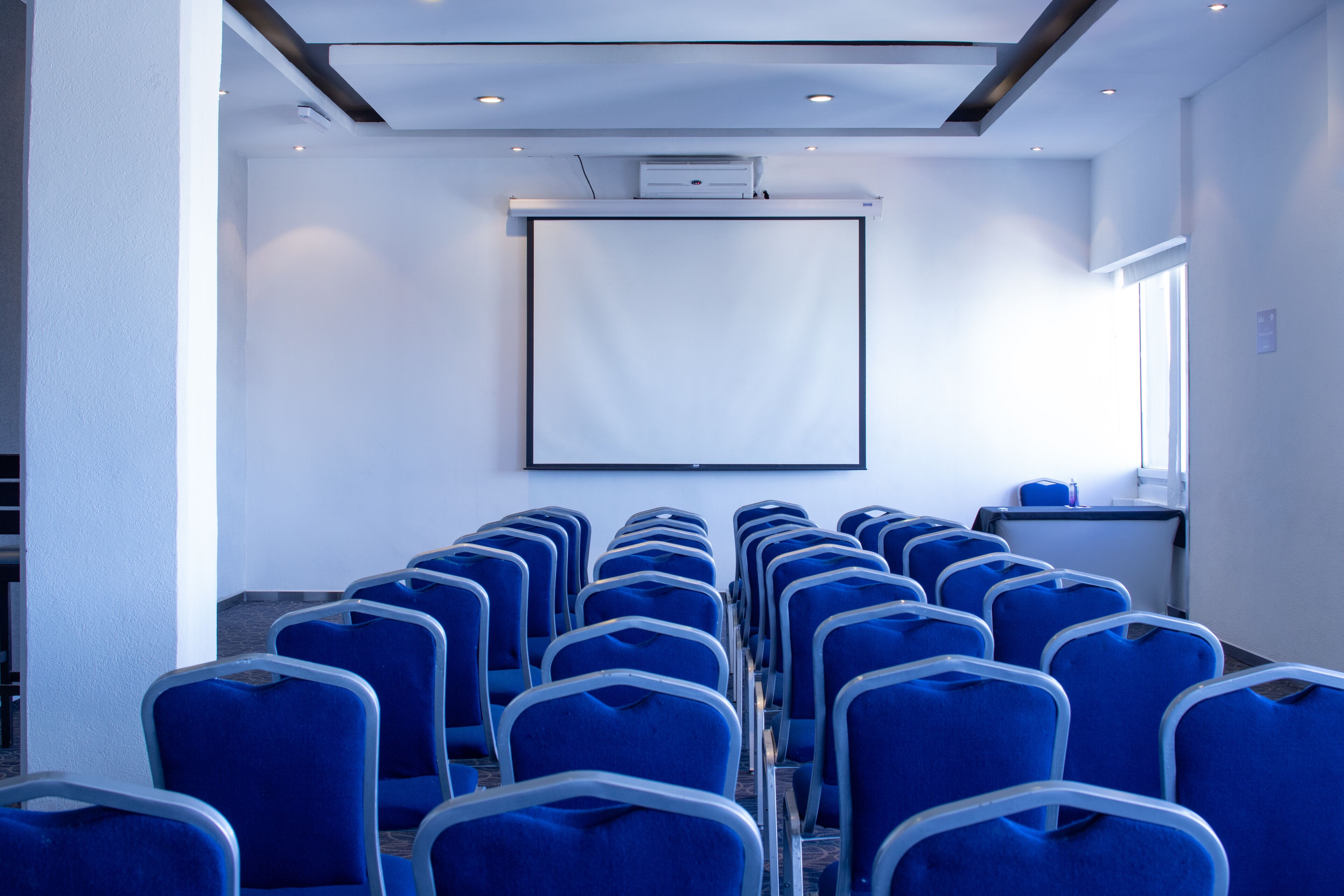 A conference room with rows of blue chairs, a projector screen, and a window providing natural light.