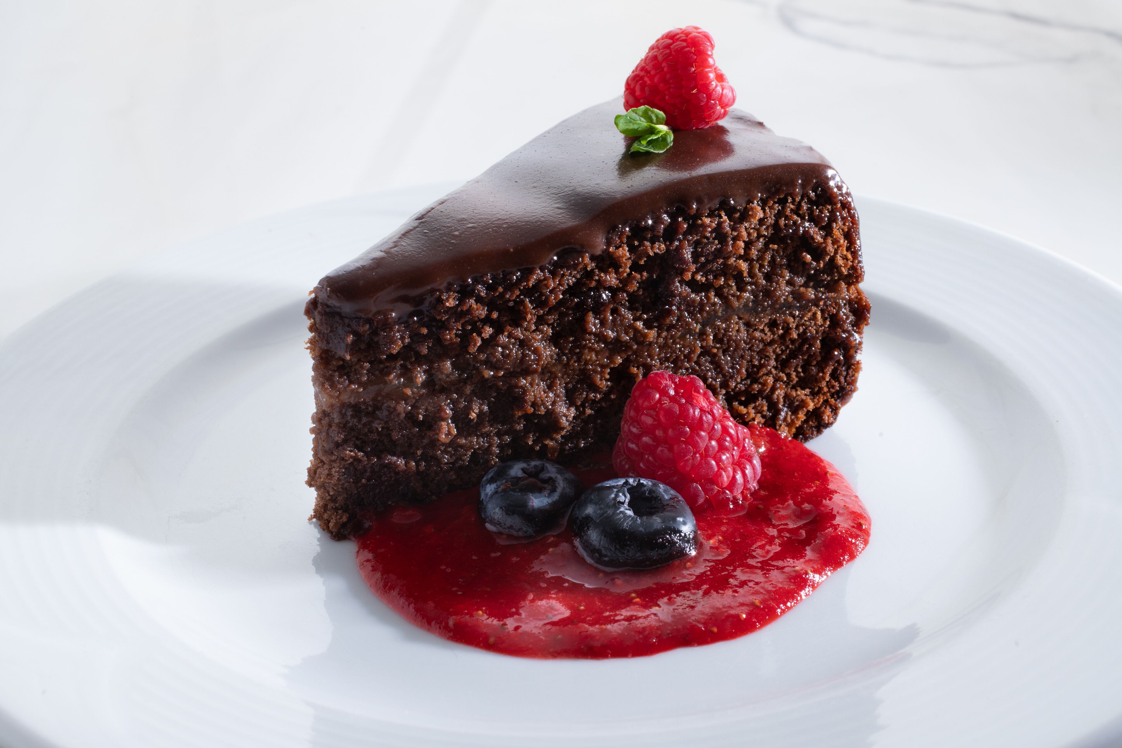 A slice of chocolate cake with chocolate glaze, a raspberry, and blueberries on a white plate.