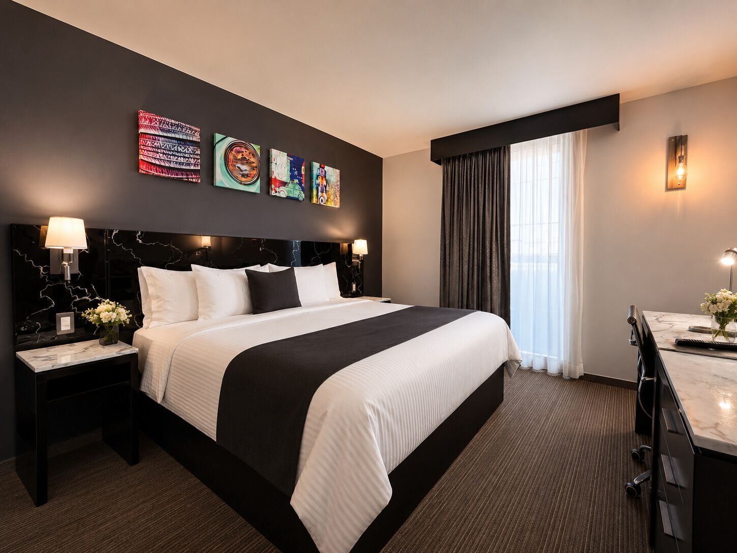 A hotel room with a large bed, a desk, and a chair. There are wall art pieces above the bed and a window with curtains.