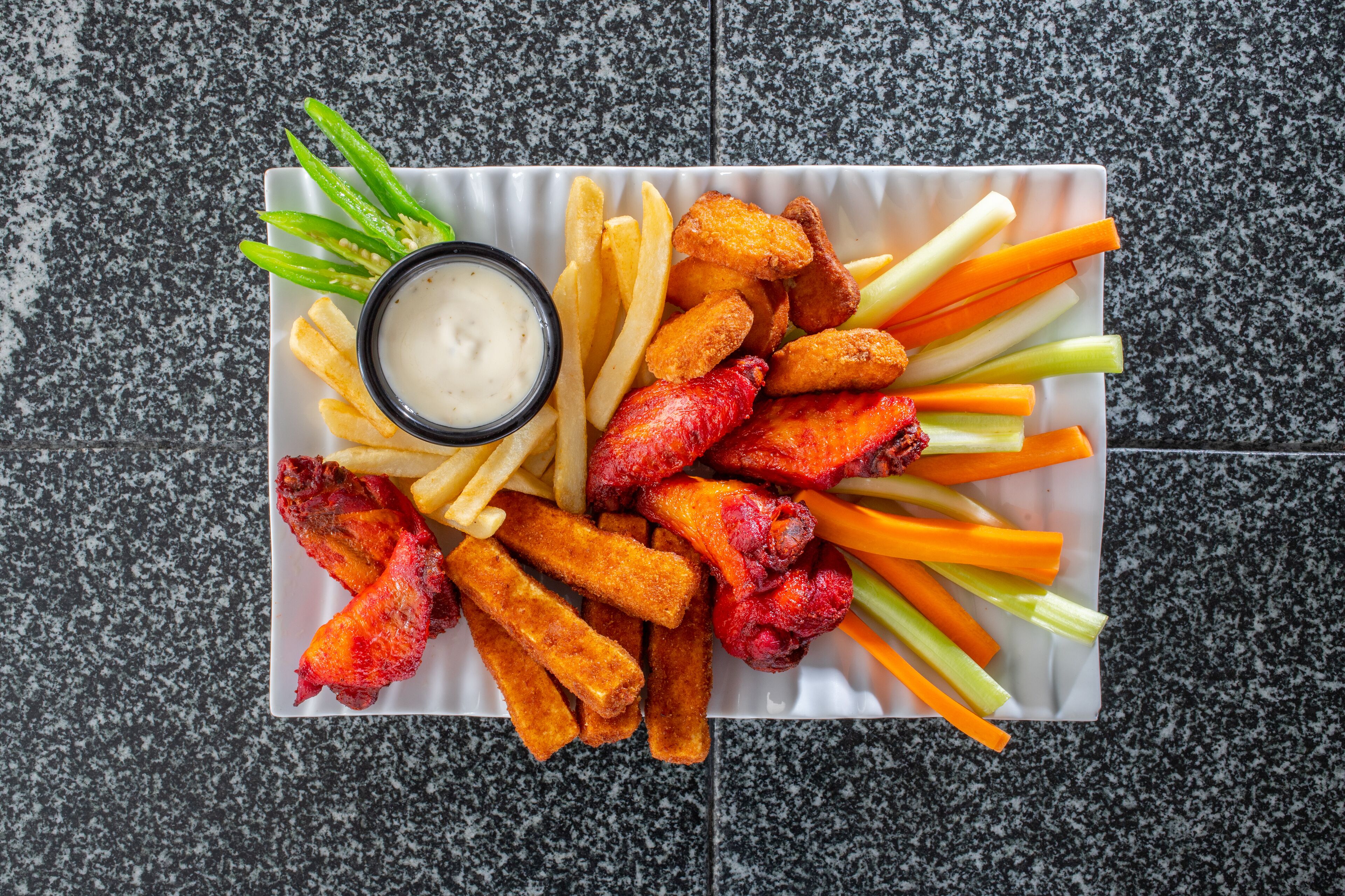 Un plato rectangular blanco con alitas de pollo fritas, papas fritas, bastones de apio y un recipiente pequeño con salsa para mojar.