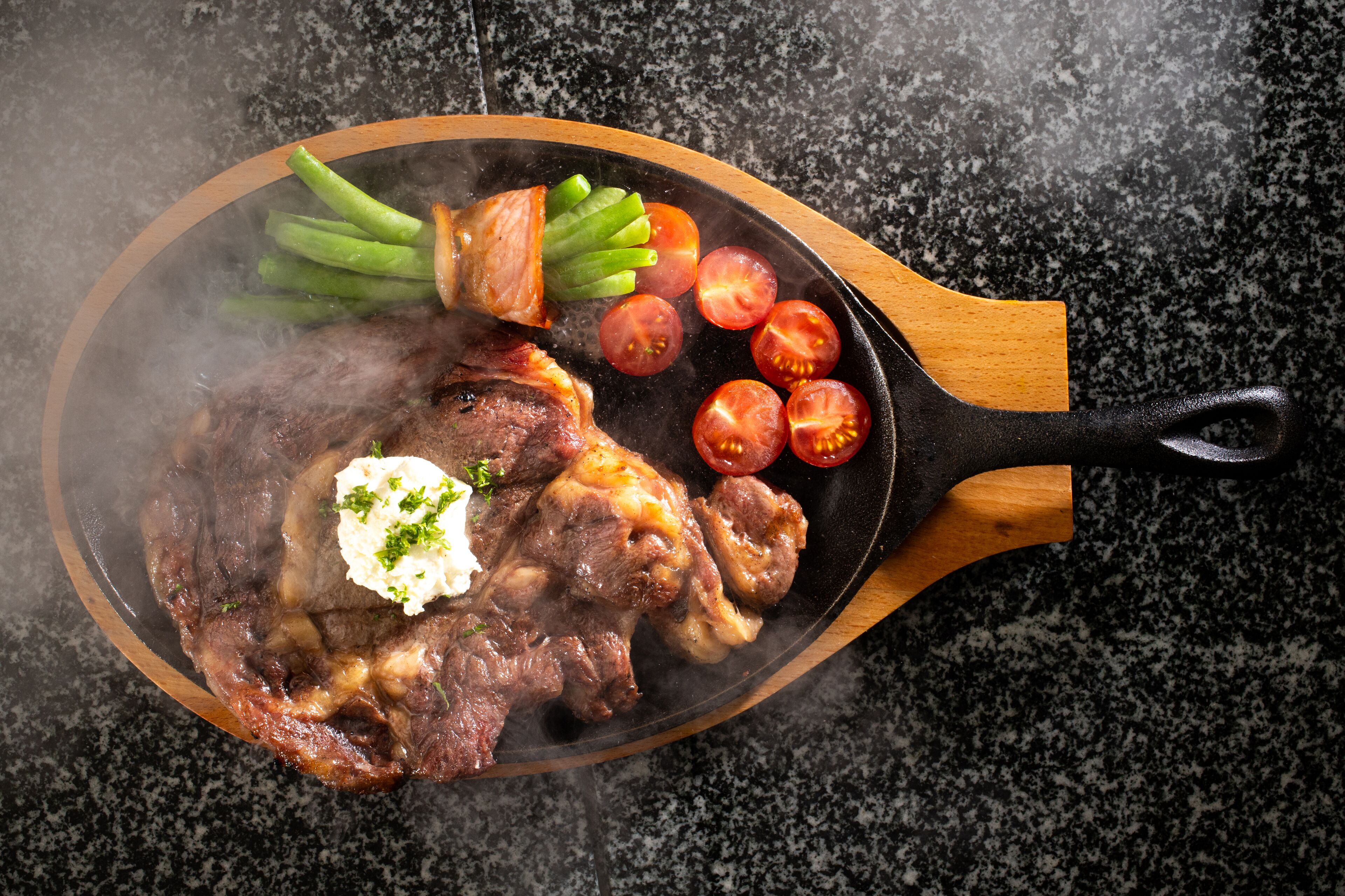 Una sartén con un bistec, judías verdes y tomates cherry sobre una tabla de madera.