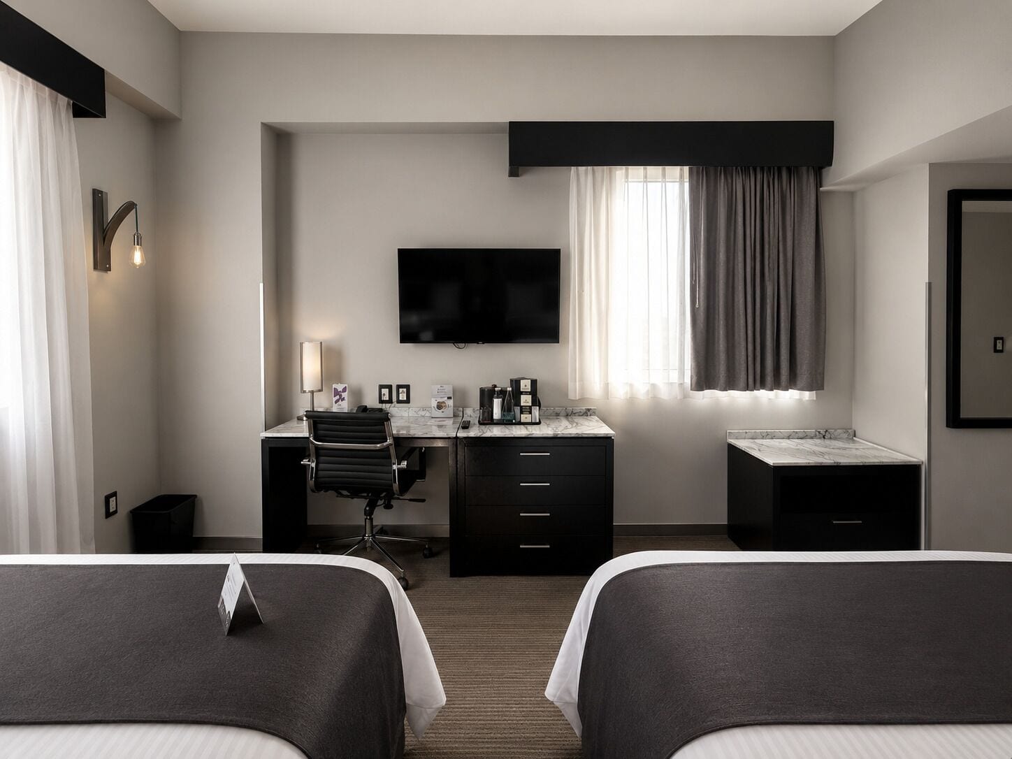 Une chambre d’hôtel avec deux lits, un bureau avec une chaise, un téléviseur à écran plat, une machine à café et une fenêtre avec des rideaux.