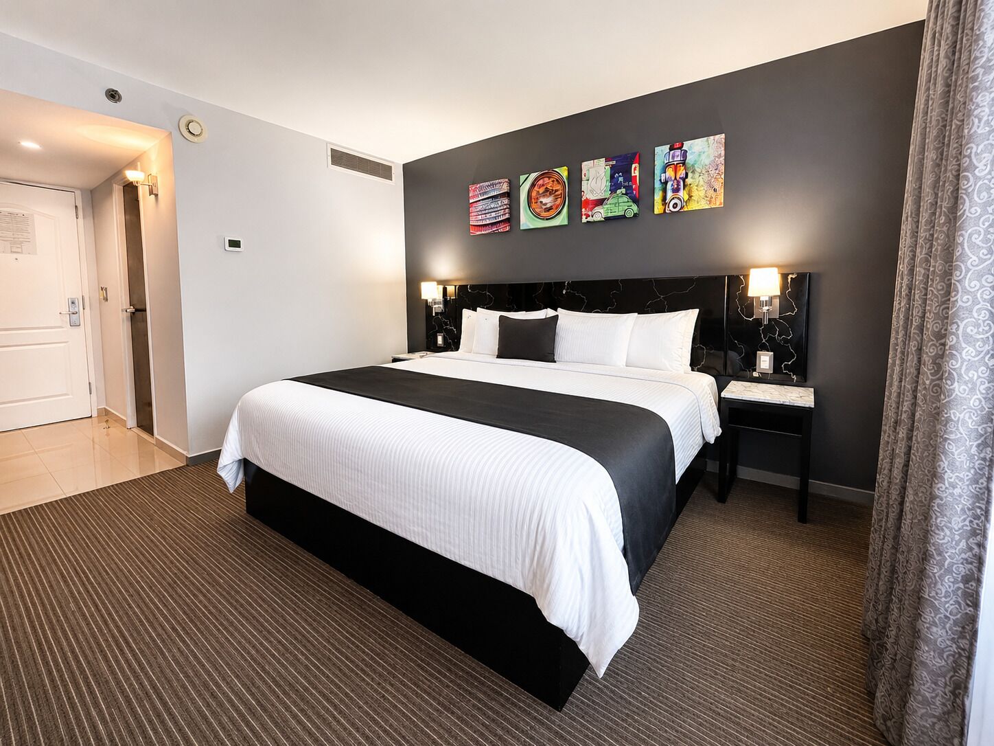 Una habitación de hotel con una cama grande, una mesita de noche, un televisor montado en la pared y una puerta.