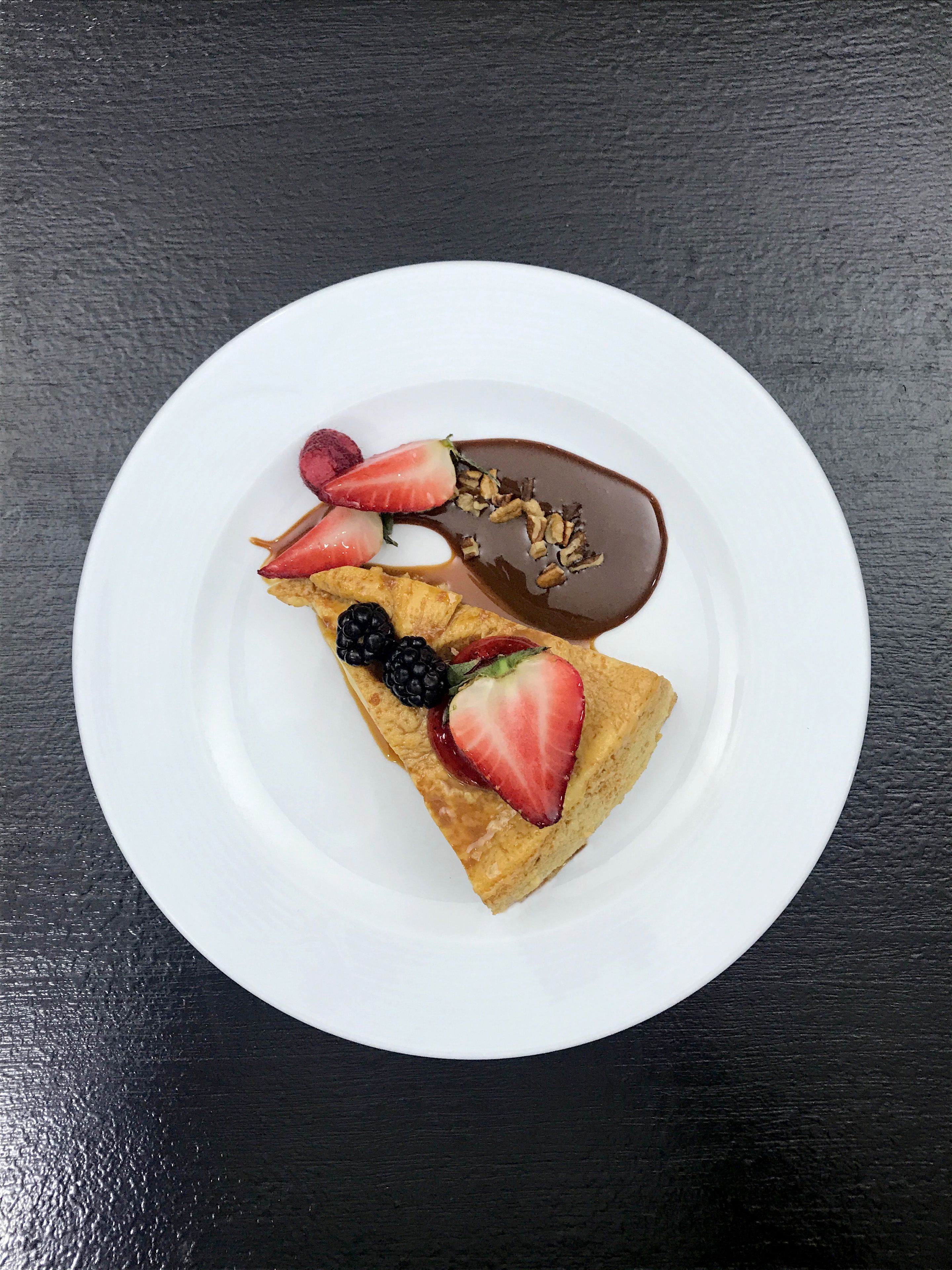A dessert on a white plate with chocolate sauce, strawberries, and blackberries on a dark surface.
