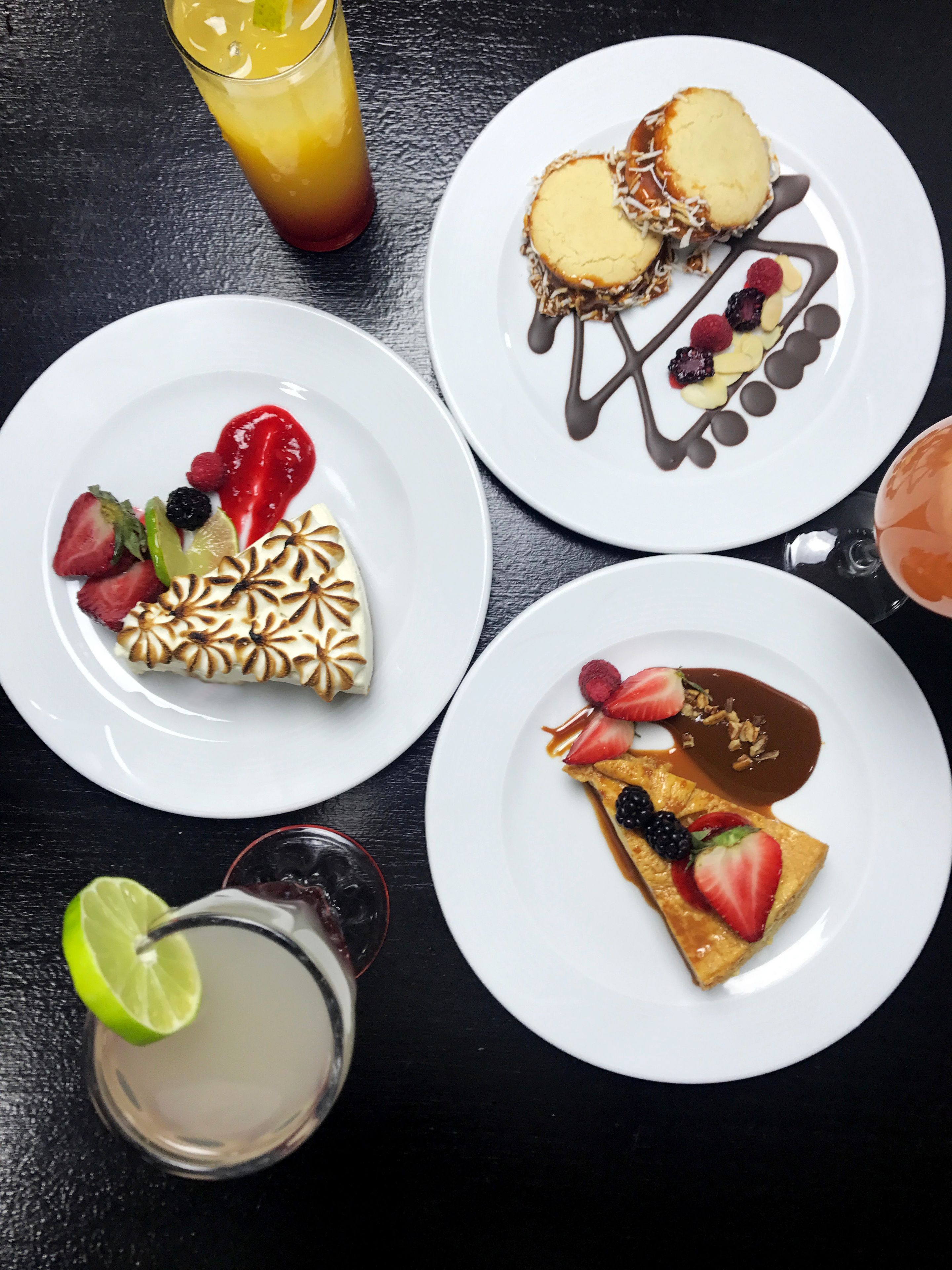 Three plates of dessert with various fruits and sauces, a glass of iced beverage, and a lime wedge on a table.