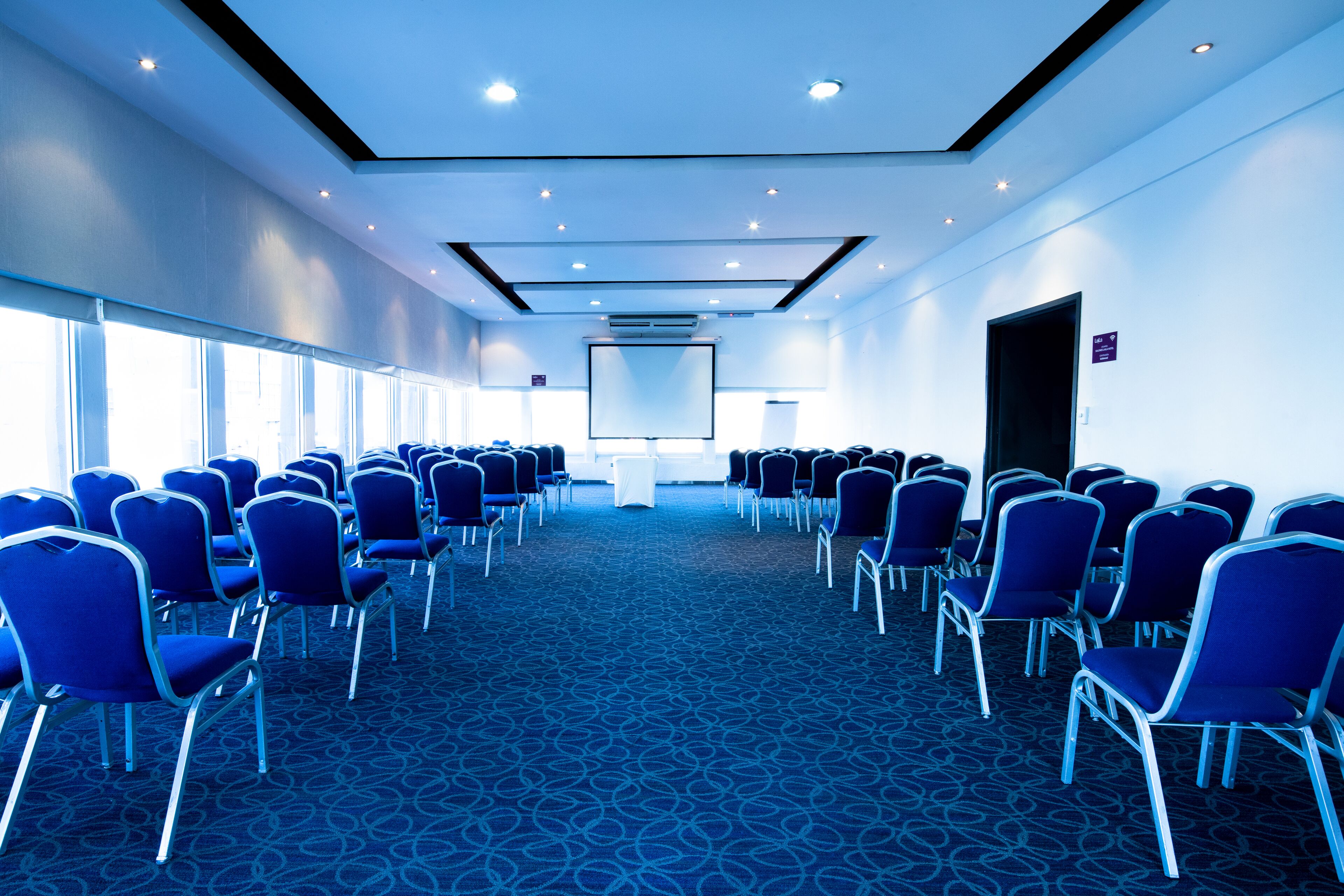 Une salle de conférence avec des rangées de chaises bleues, un grand écran à l’avant et un pupitre blanc.