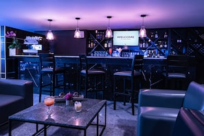 Zona de bar con una pantalla de bienvenida, taburetes, barra y un área de asientos con una mesa y sillas.