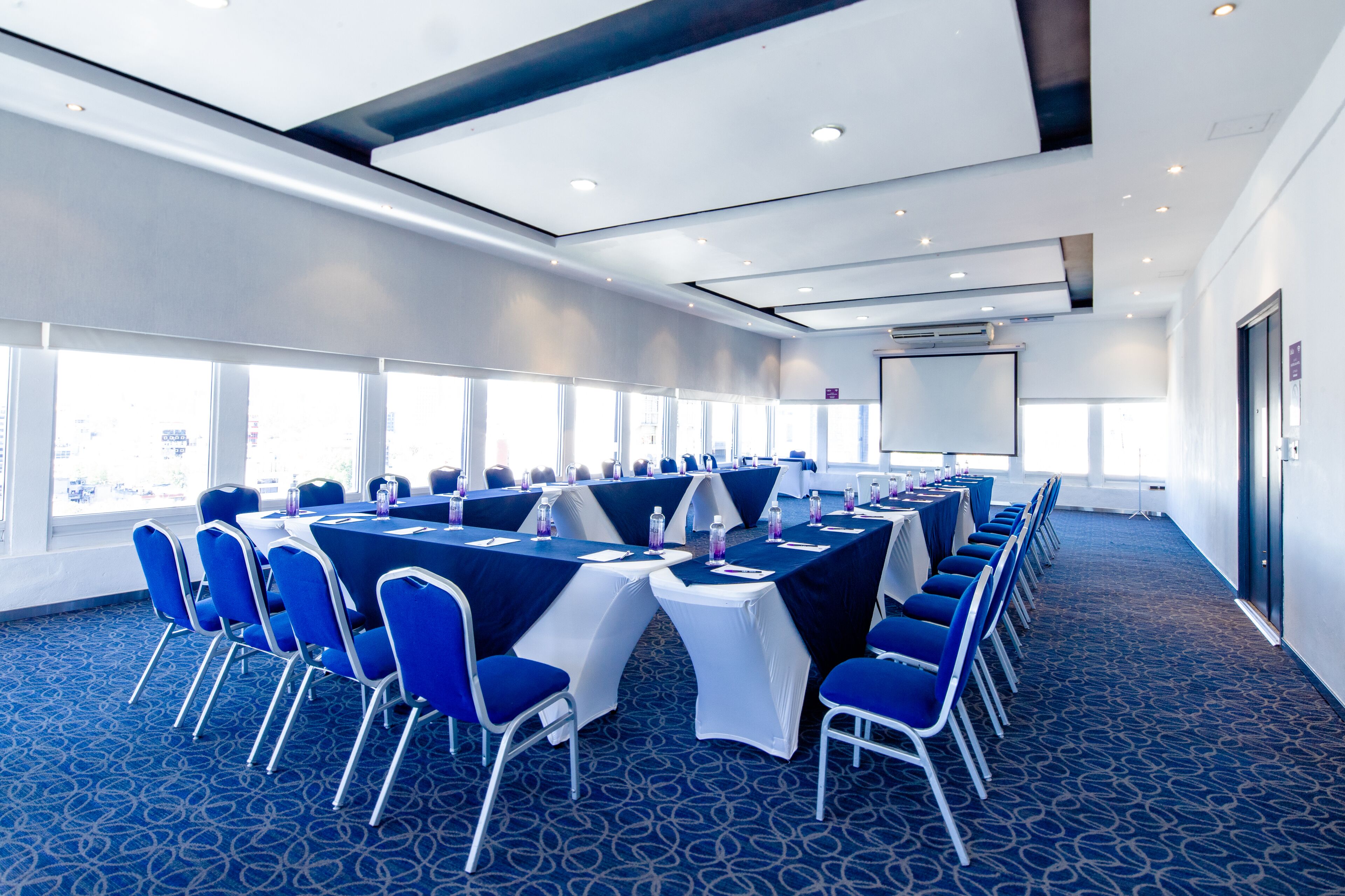 A conference room with blue chairs and tables, large windows, and a projector screen.