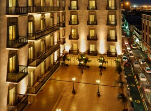 Um hotel com vários andares, varandas, um pátio com plantas em vasos e vista para a rua com carros.