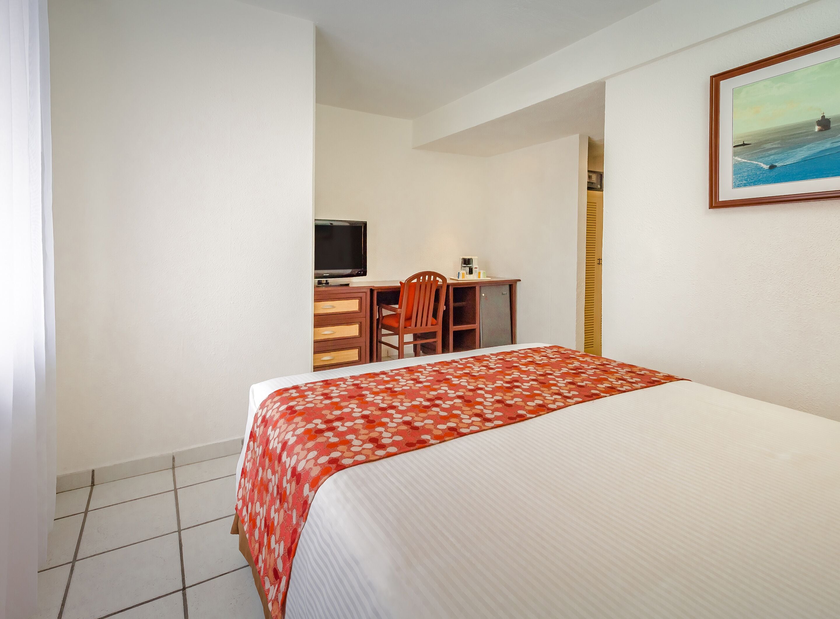 Habitación de hotel con cama, escritorio, silla, televisor y un cuadro en la pared.