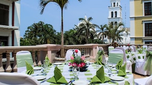 A dining area with tables set for a meal, palm trees, and a white building with a bell tower in the background. - Veracruz Centro Histórico (Veracruz)