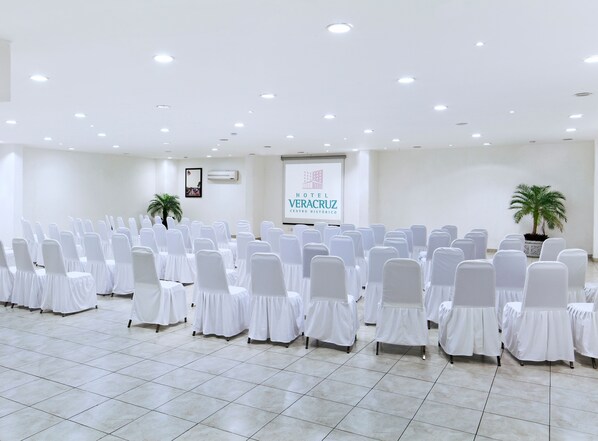 A hotel conference room with rows of white chairs, a projector screen, and two potted plants.