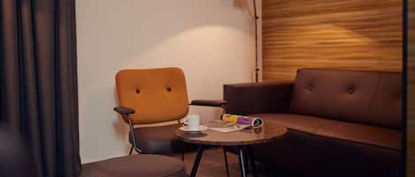 Una habitación moderna con un sofá de cuero marrón, una silla de madera, una mesita redonda pequeña con una taza y una revista, y una lámpara de pie.