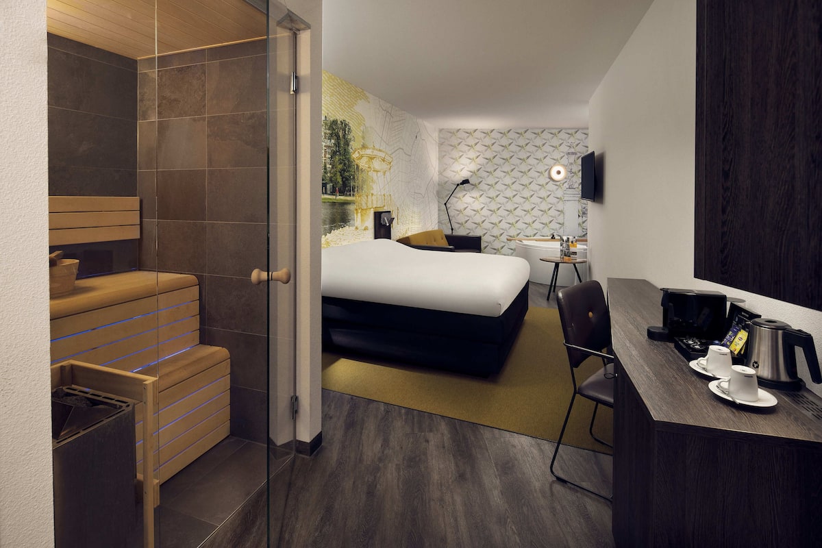 A modern hotel room with a bed, a desk with a coffee maker, a chair, and a shower area.