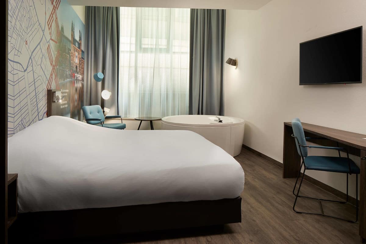 A hotel room with a large bed, a desk, a chair, a round bathtub, and a TV on the wall.