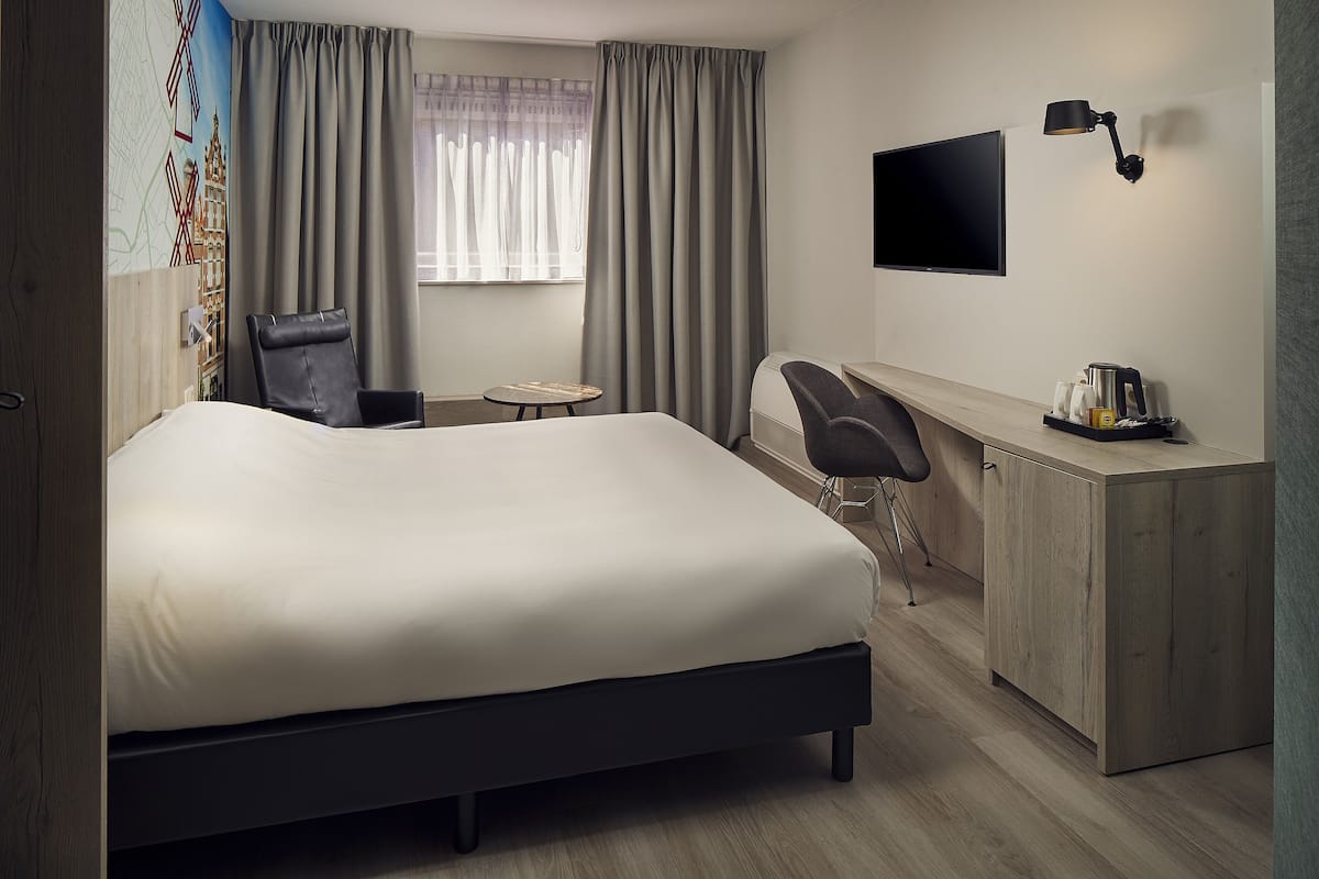 A hotel room with a large bed, a desk, a chair, a TV, and a window with curtains.