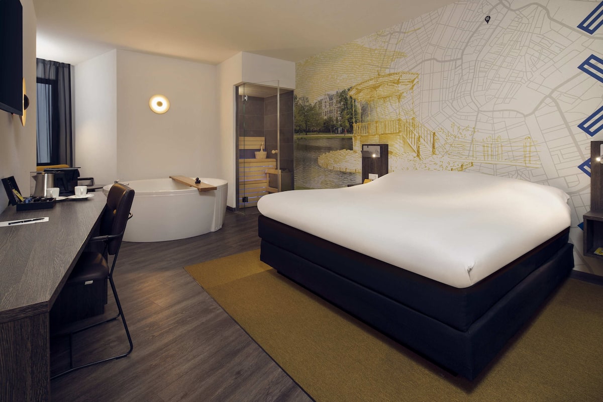 A modern hotel room with a large bed, a desk with a chair, a bathtub, and a map mural on the wall.