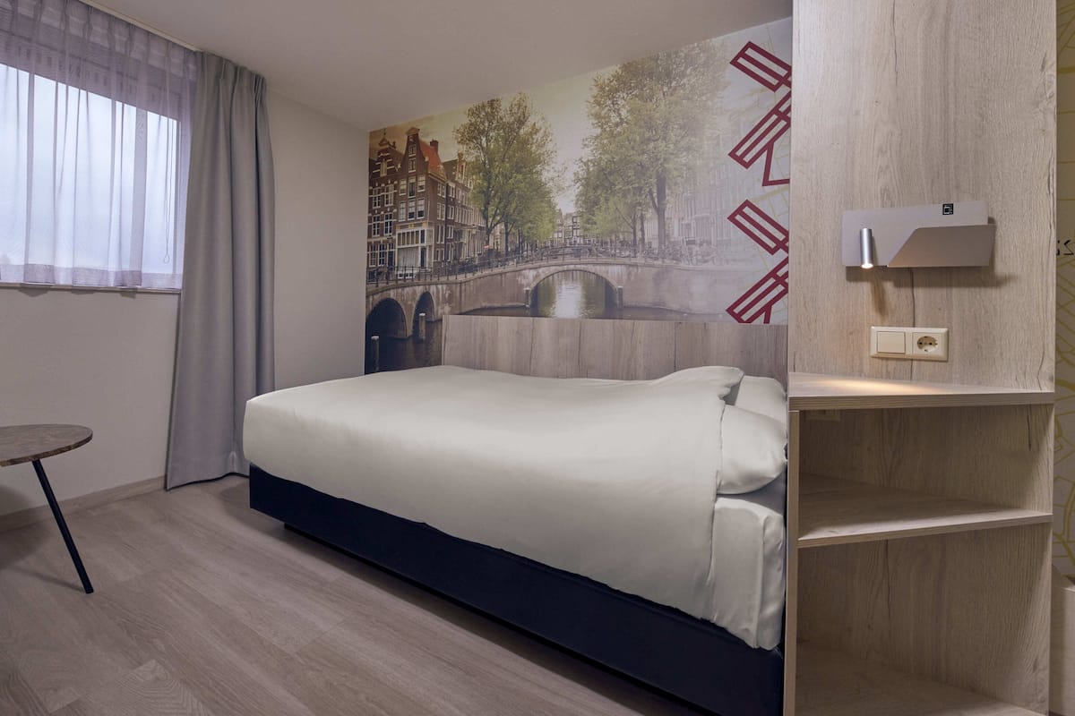 A hotel room with a bed, a small table, a chair, a window with curtains, a wall-mounted light, and a wooden shelf.