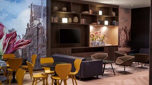 Una sala moderna con un televisor de pantalla plana grande, estanterías de madera con libros y objetos decorativos, un sofá, sillas amarillas y un gran mural floral en la pared.